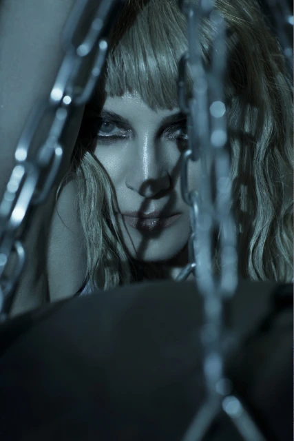 Close-up portrait of a fair-skinned woman with blond hair, styled with blunt bangs, looking at the camera through blurred metal chains.