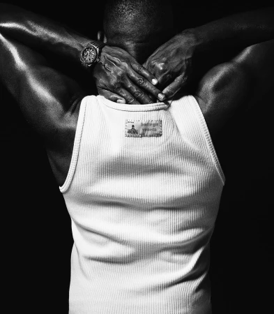 Black and white portrait of a man from behind wearing a ribbed white tank top with his hands behind his neck.