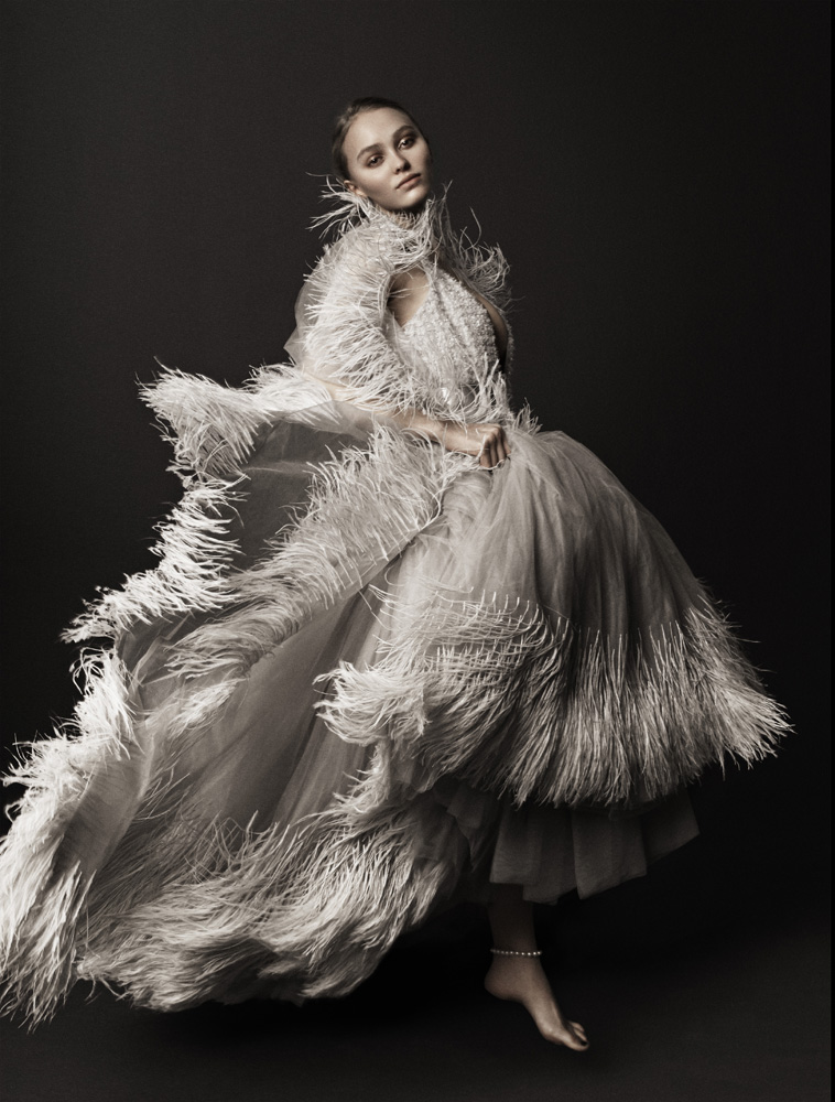Elegant woman in feathered gown poses in studio, creating an editorial-style fashion portrait with a dark background and soft, diffused lighting.