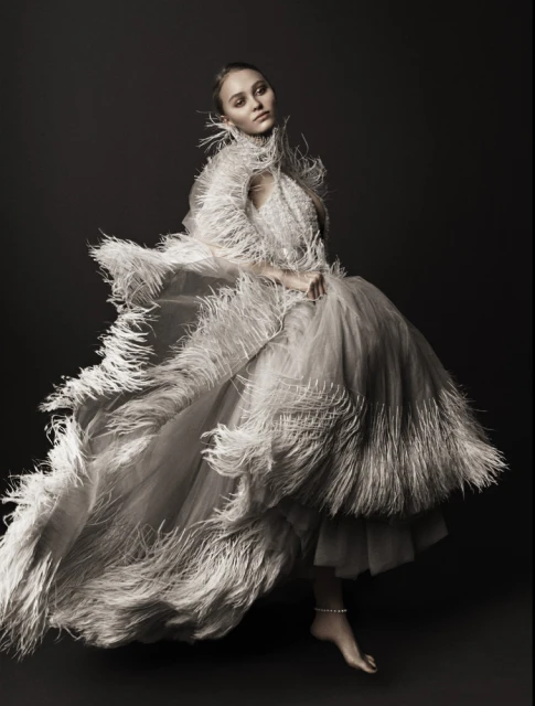 Elegant woman in feathered gown poses in studio, creating an editorial-style fashion portrait with a dark background and soft, diffused lighting.
