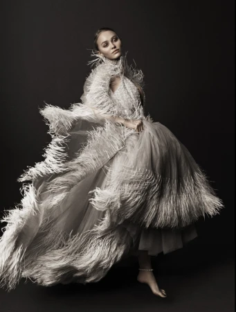 Elegant woman in feathered gown poses in studio, creating an editorial-style fashion portrait with a dark background and soft, diffused lighting.
