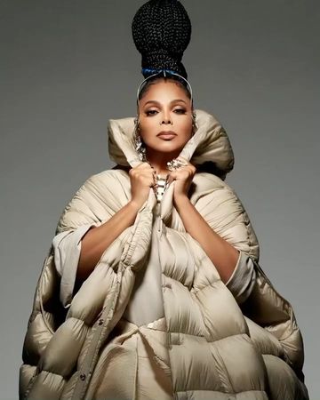 Adult woman wearing a large beige puffer coat with hair in braids and silver earrings against a grey background in a studio shot.