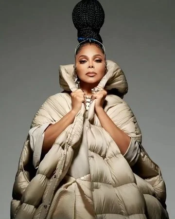 Adult woman wearing a large beige puffer coat with hair in braids and silver earrings against a grey background in a studio shot.
