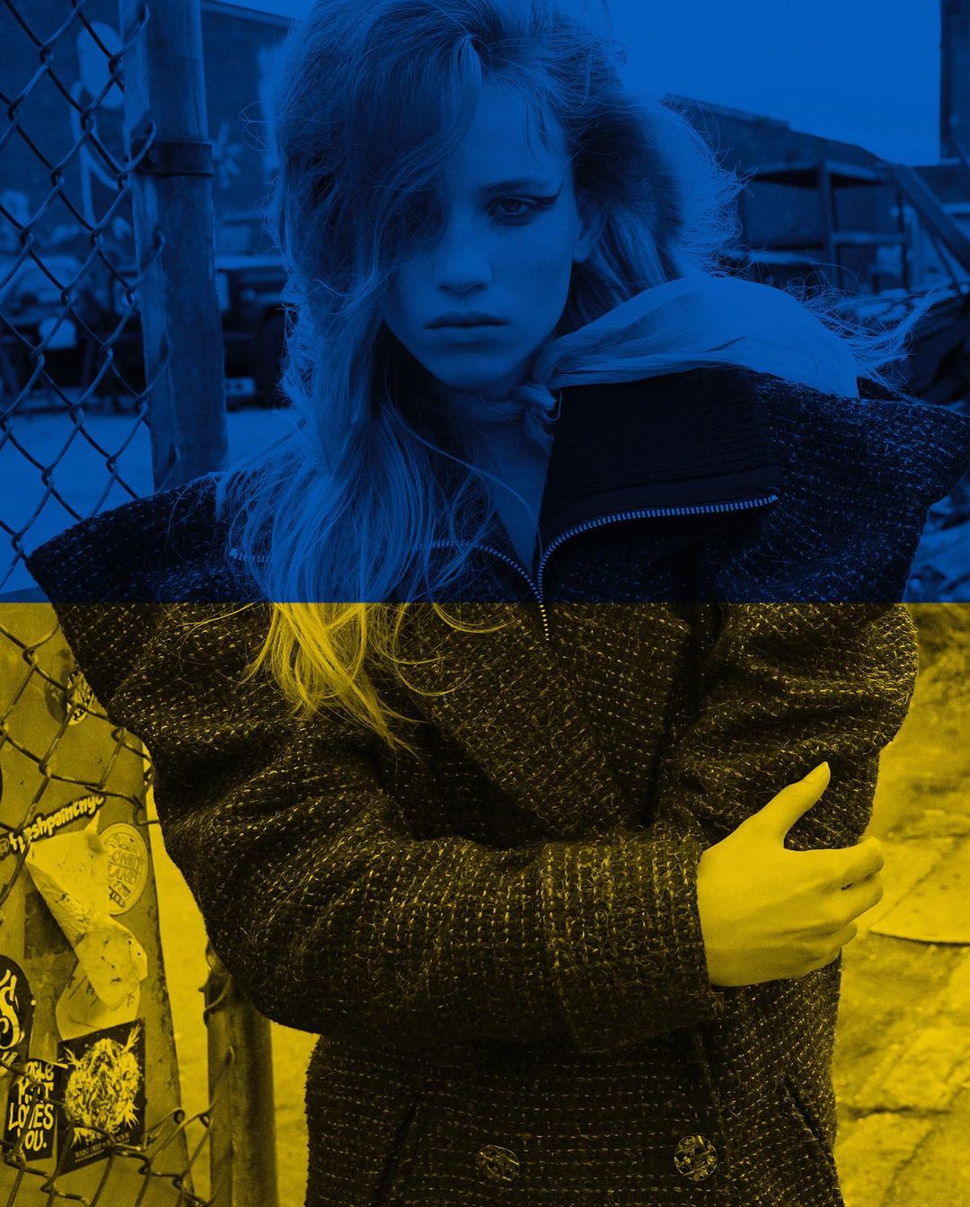 Fashionable woman in coat overlaid with Ukraine flag colors, standing outdoors against a metal fence in an urban environment.