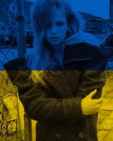 Fashionable woman in coat overlaid with Ukraine flag colors, standing outdoors against a metal fence in an urban environment.