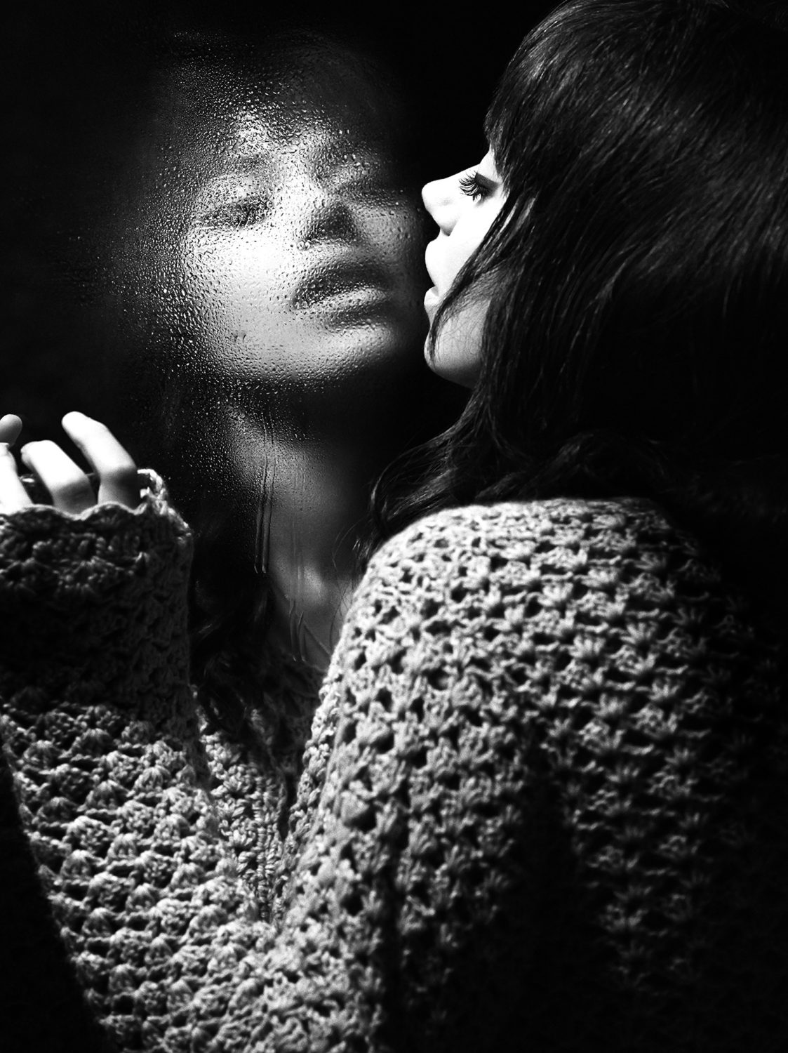 Black and white portrait of a woman looking through a rain-streaked window with a textured knitted sweater.