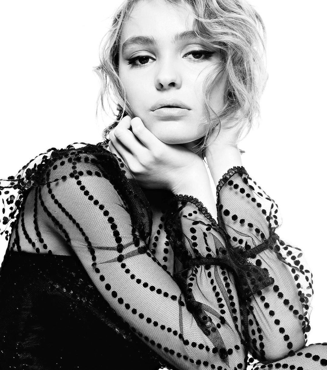Black-and-white portrait of a young woman with wavy blonde hair, looking at the camera in a patterned top against a white backdrop.