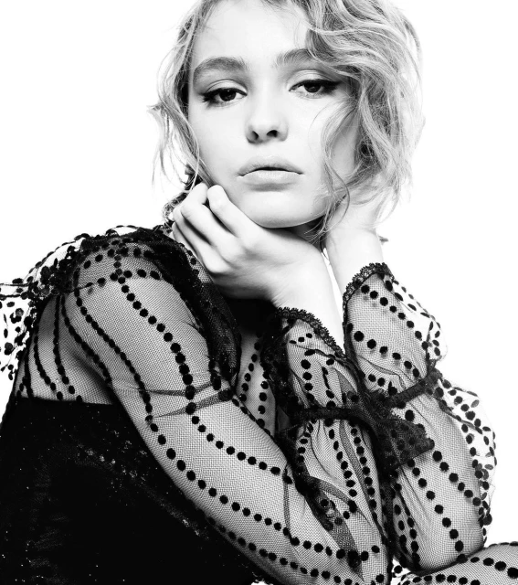 Black-and-white portrait of a young woman with wavy blonde hair, looking at the camera in a patterned top against a white backdrop.