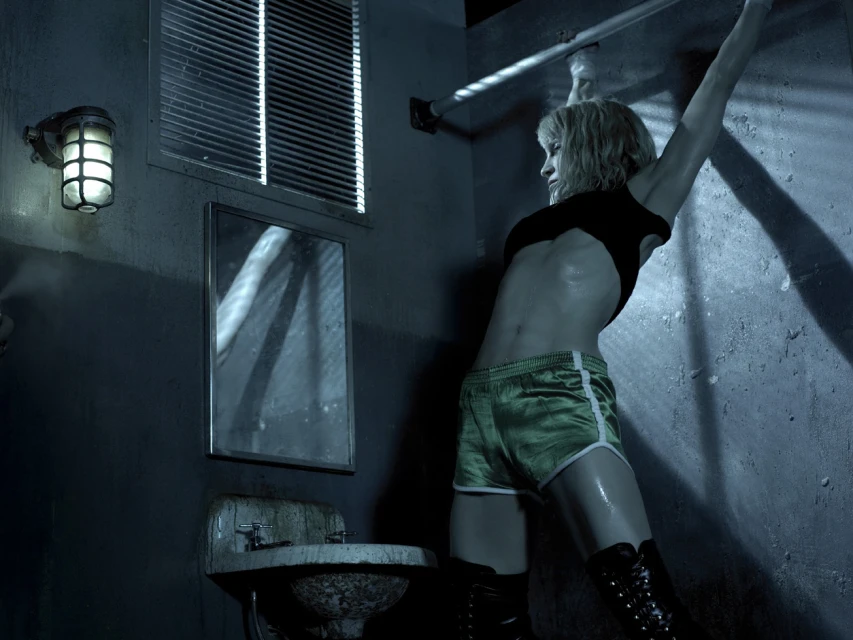Athletic woman in green shorts and black boots leans against a wall in a grungy, dimly lit room with a sink, mirror, and barred window.