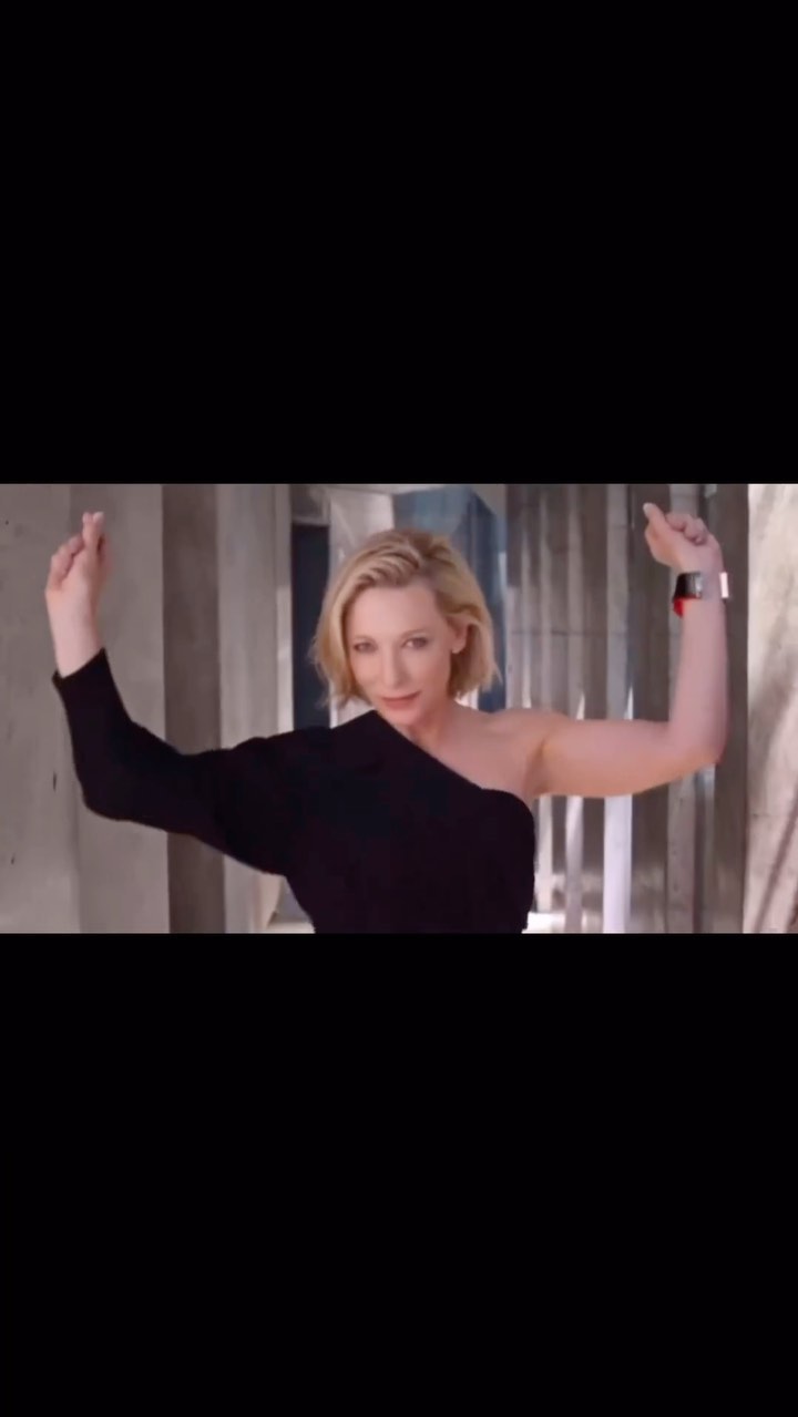 Blonde woman with short hair wearing a one-shoulder black dress poses in a modern architectural space, arms raised.
