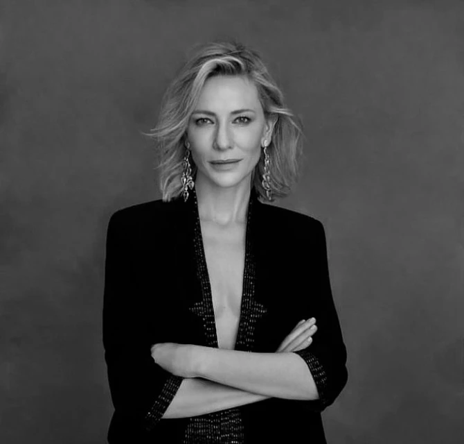 Black-and-white portrait of a blonde woman in a black jacket with arms crossed, wearing chandelier earrings against a grey backdrop.