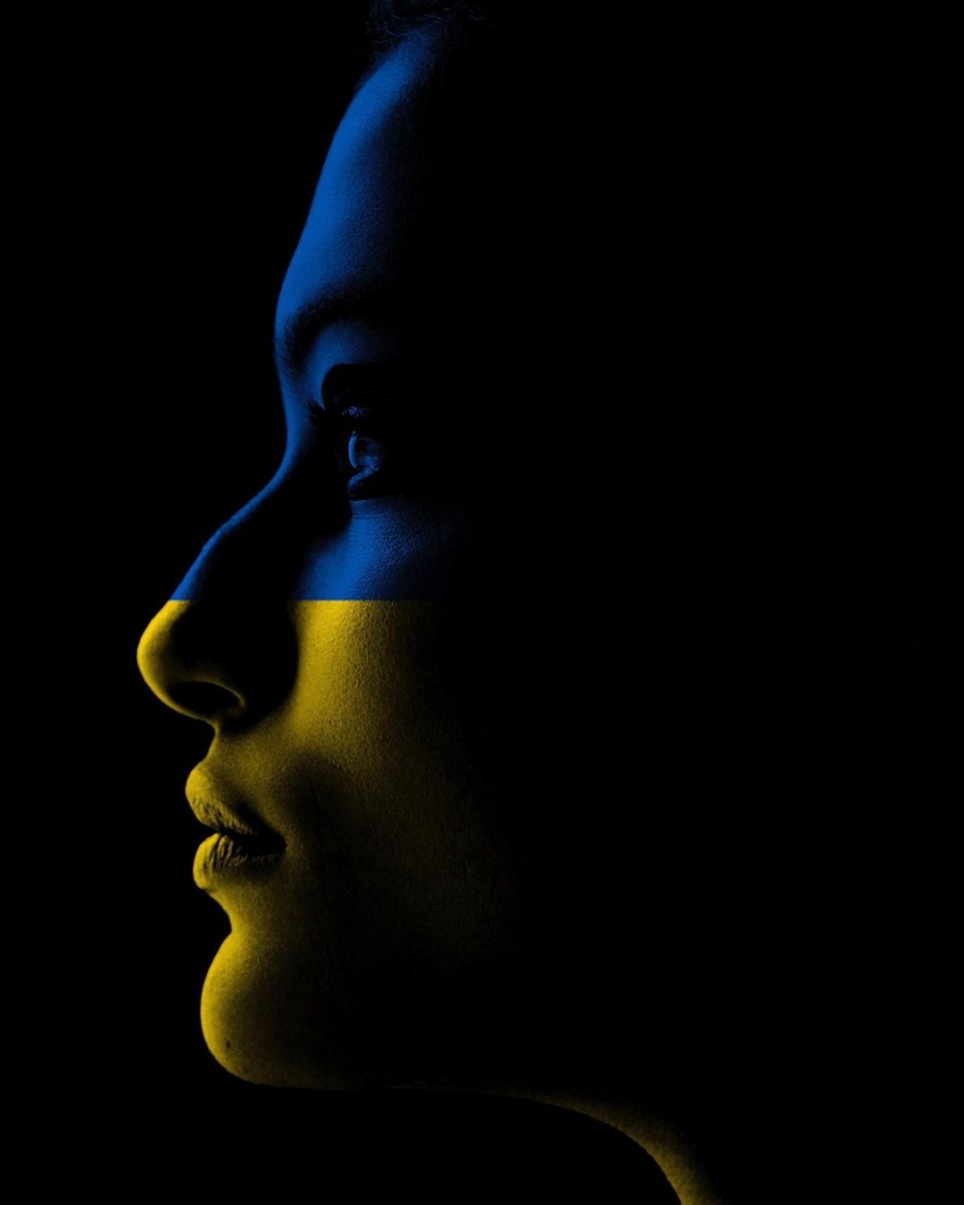 Profile view of a woman's face with the colors of the Ukrainian flag against a black background in a dramatic, artistic style.