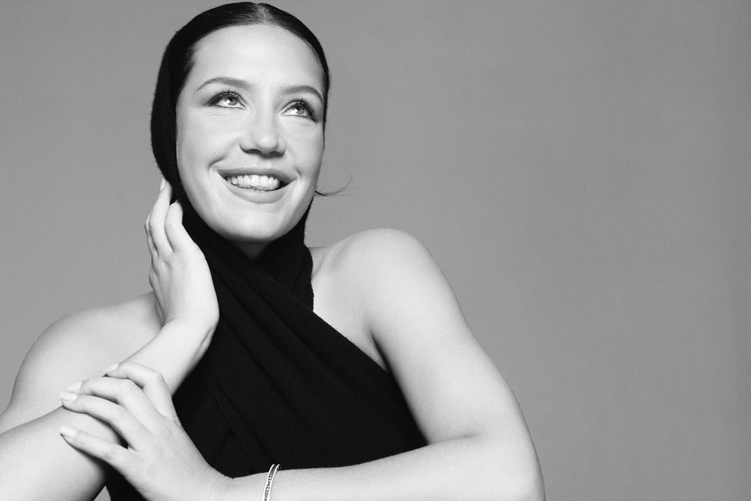 Black and white portrait of a smiling woman with hands near her face, wearing a dark head covering and top, set against a grey background.