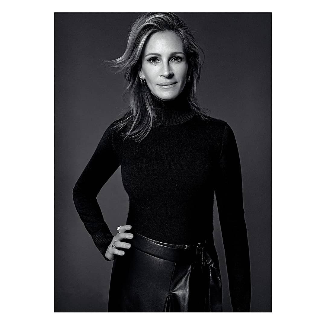 Black and white portrait of adult female with long brown hair wearing a black turtleneck, skirt and ring, set against a grey backdrop.