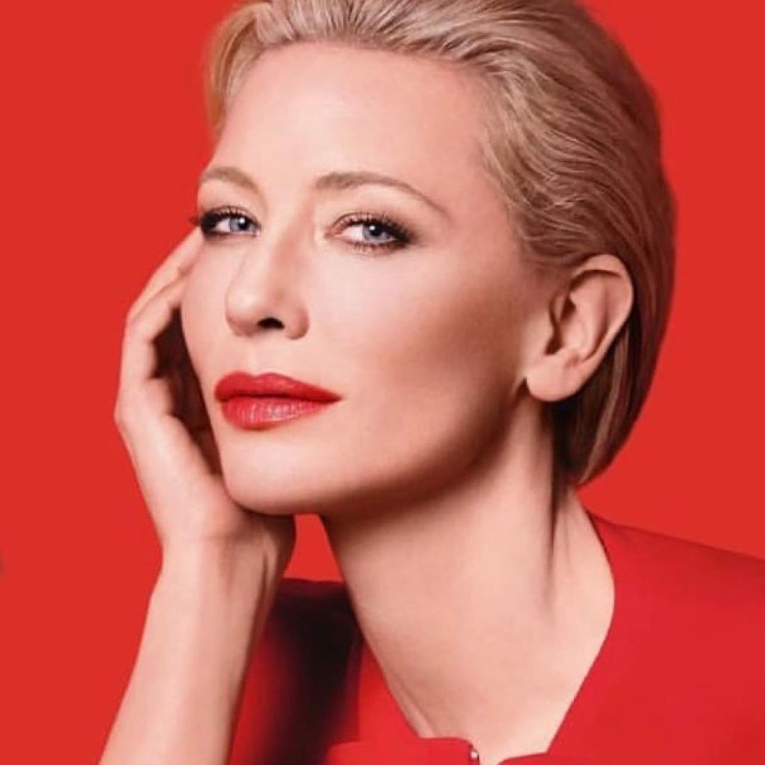 Close-up portrait of a blonde woman with red lipstick, gazing calmly at the camera against a solid red background in a beauty shot.