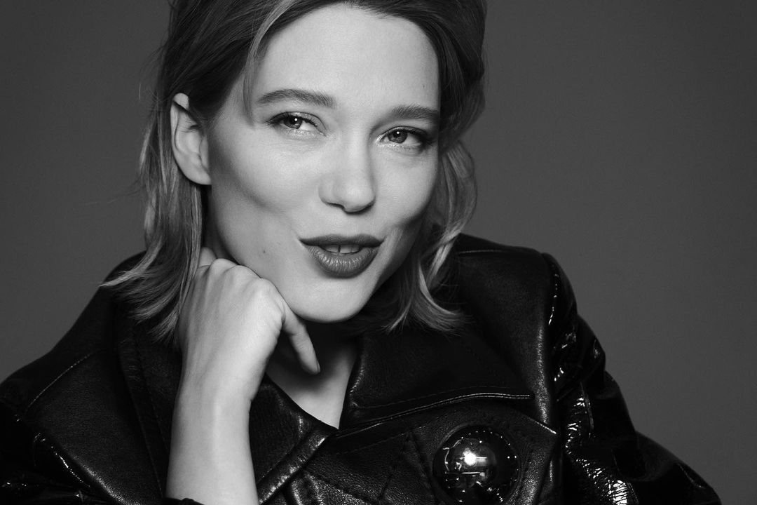 Black and white portrait of adult female with blonde hair, wearing a shiny black jacket with a silver embellishment, indoors on grey background.