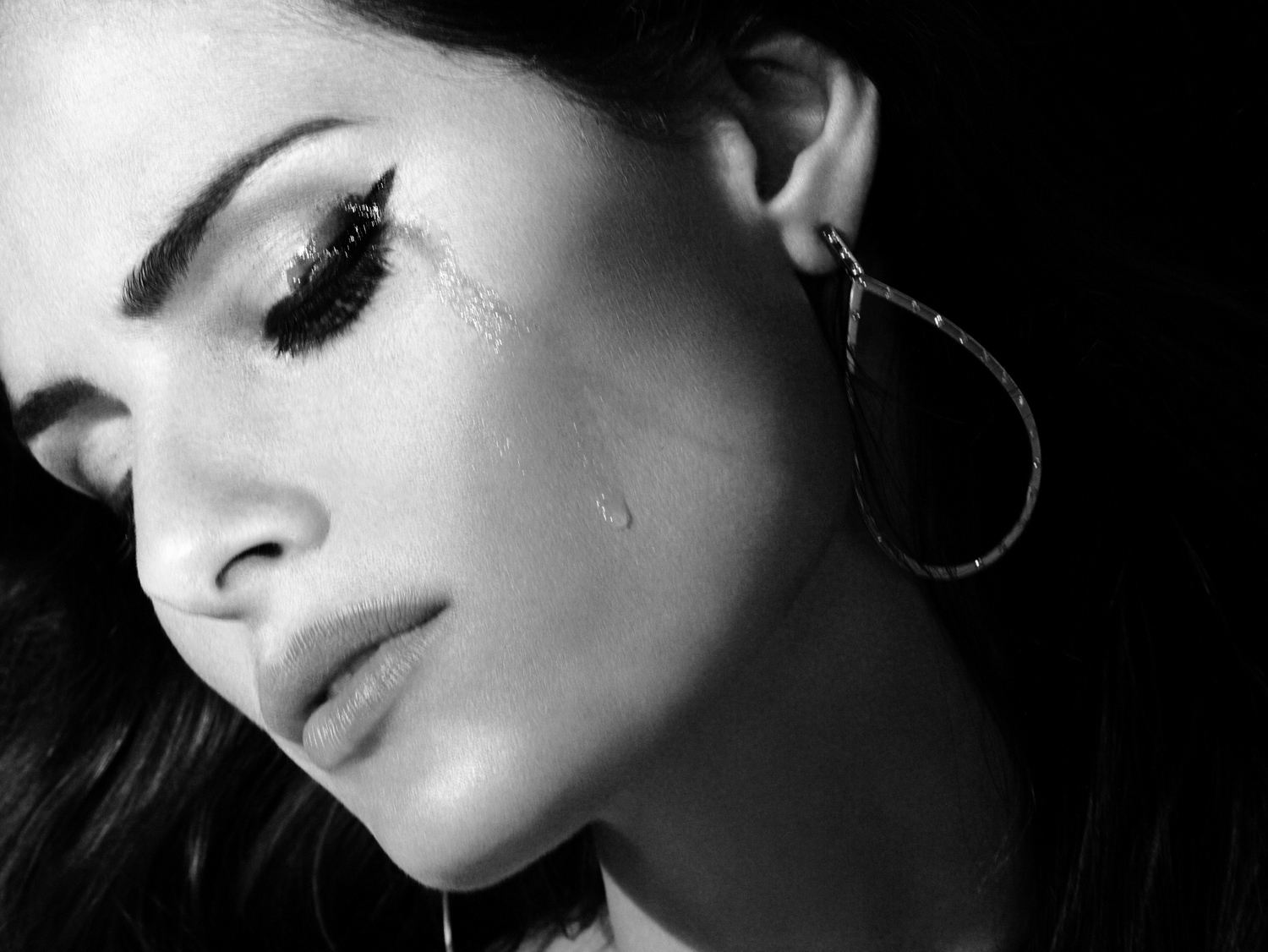 Black and white close-up of a woman with dramatic eye makeup and teardrop on her cheek, highlighting beauty and emotional expression.