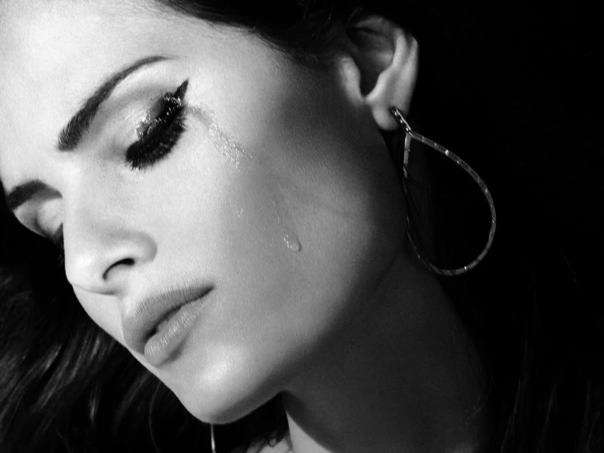 Black and white close-up of a woman with dramatic eye makeup and teardrop on her cheek, highlighting beauty and emotional expression.