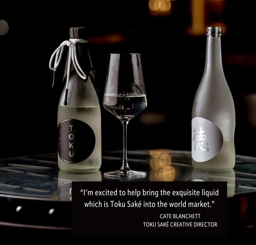Toku Sake bottles and a stemmed wine glass filled with sake are displayed on a reflective surface against a dark background.