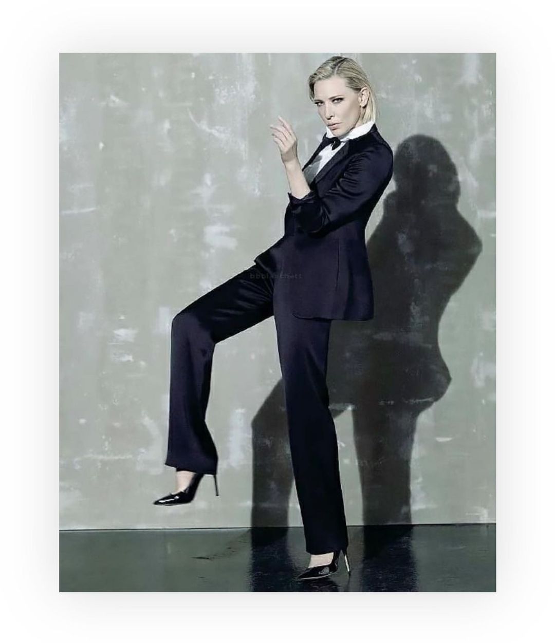 Elegant woman in a black suit and heels strikes a confident pose against a mottled grey backdrop, blending fashion and sophistication.