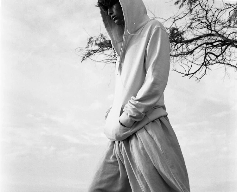Monochrome portrait of a slender adult male in a hoodie and loose pants outdoors with a cloudy sky and bare tree branches.
