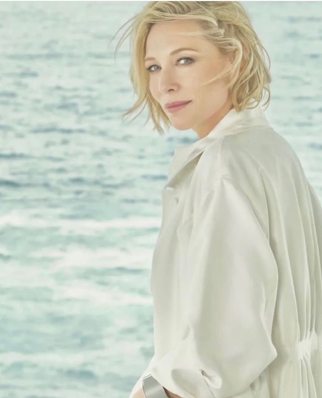Close-up shot of a blonde adult female model in a cream-colored jacket with a serene expression, set against a blue water background.