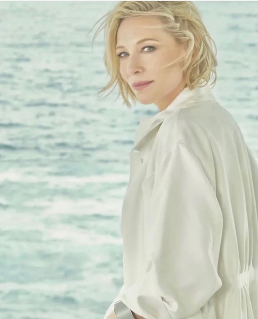 Close-up shot of a blonde adult female model in a cream-colored jacket with a serene expression, set against a blue water background.