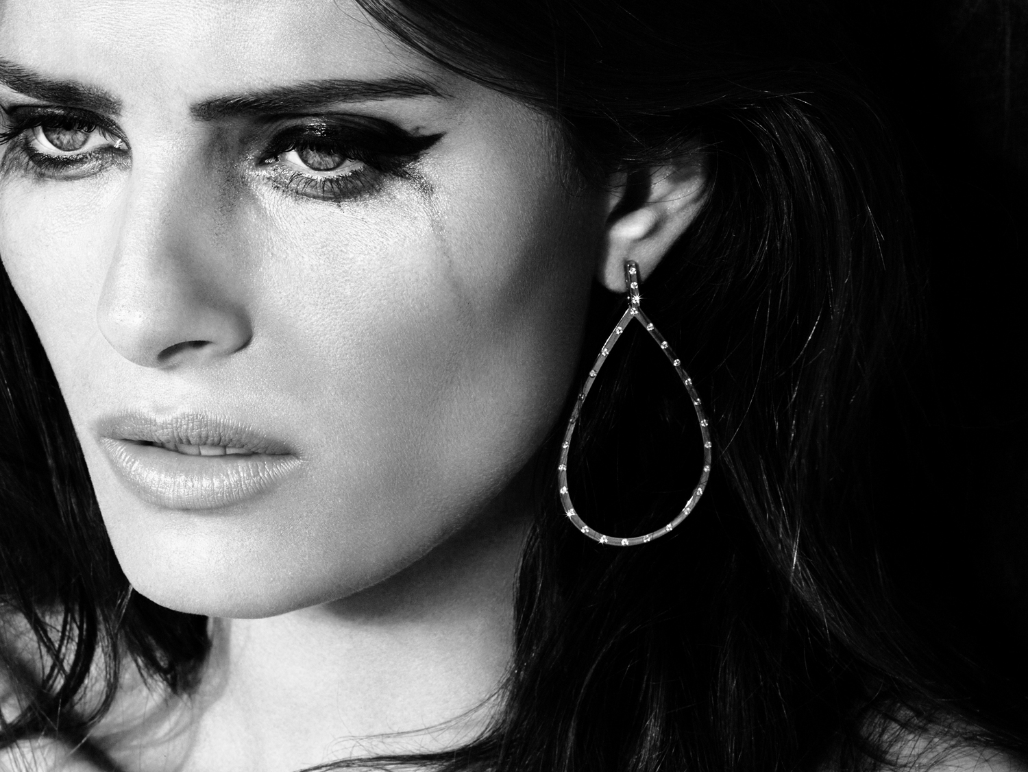 Black and white portrait of a woman with dramatic makeup and teardrop earrings, featuring a close-up studio shot with a dark background.
