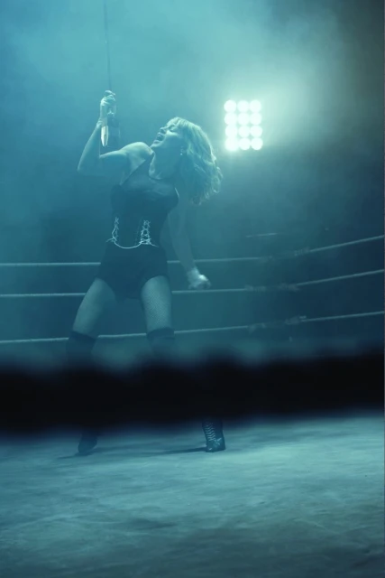 Female boxer strikes a pose in a boxing ring with dramatic lighting and smoky background. She is wearing a black outfit and knee pads.