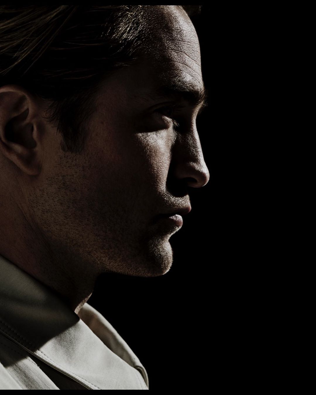 Portrait of adult man with dark hair in profile wearing a jacket, dramatically lit against a black background in a studio shot.