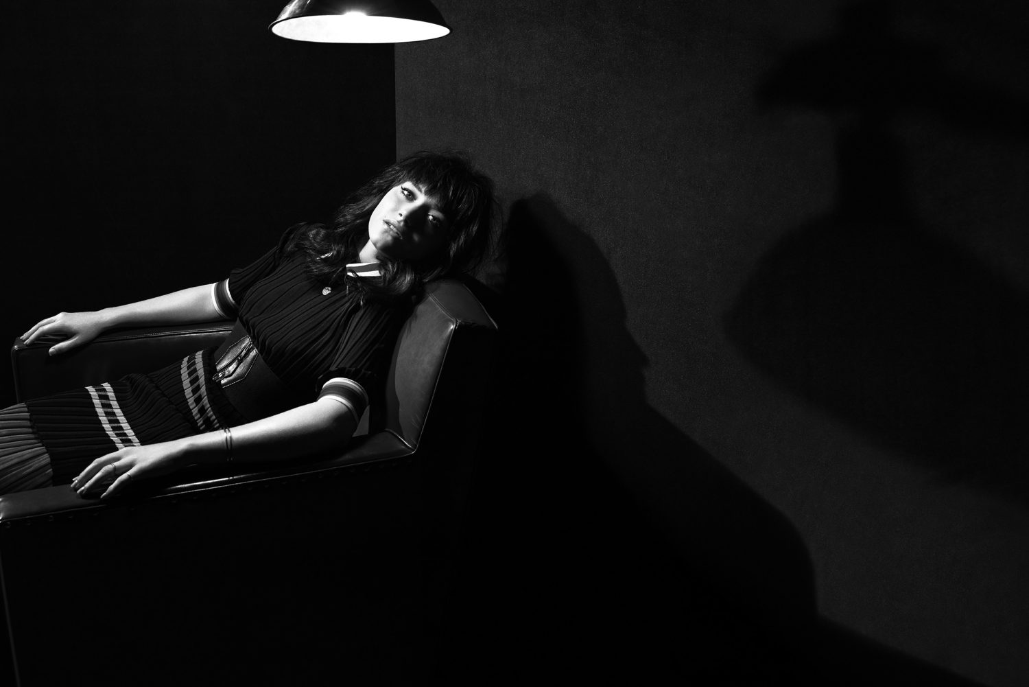 Black and white portrait of a brunette woman lounging in a chair, under a hanging light against a dark background with her shadow.