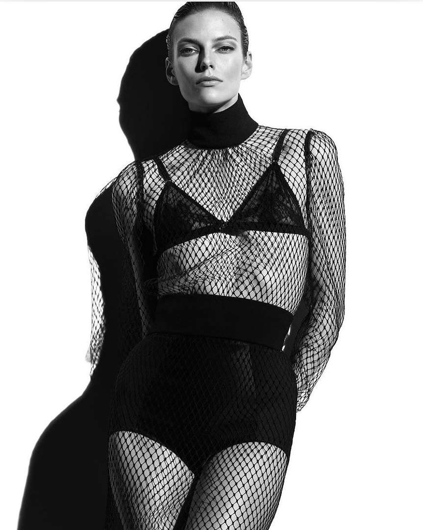 Monochrome fashion shot of a woman in fishnet top and shorts standing in front of her shadow against a white background in studio lighting.