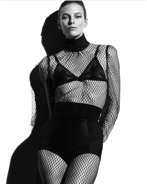 Monochrome fashion shot of a woman in fishnet top and shorts standing in front of her shadow against a white background in studio lighting.
