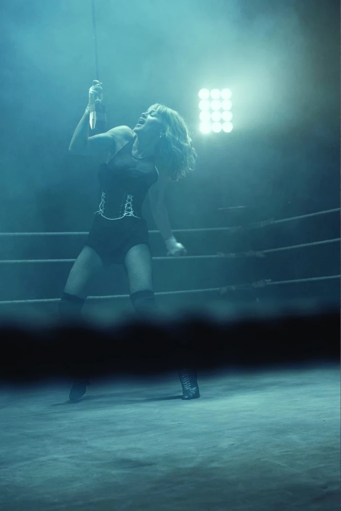 Blonde woman in dark athletic bodysuit standing inside a boxing ring under stage lights amidst haze.