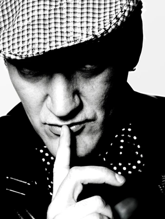Close-up portrait of a man wearing a houndstooth flat cap and polka-dot shirt with his finger to his lips in a monochrome photograph