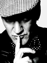 Close-up portrait of a man wearing a houndstooth flat cap and polka-dot shirt with his finger to his lips in a monochrome photograph