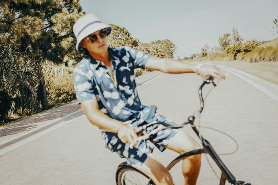 Adult male in patterned shirt and bucket hat rides a bicycle on a paved road lined with green foliage on a sunny day.