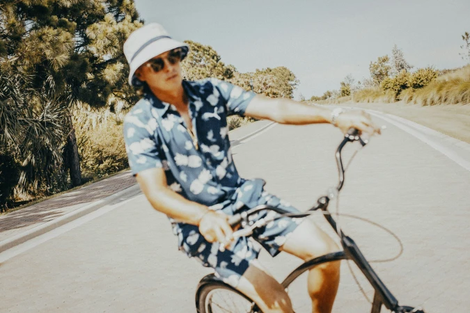 Adult male in patterned shirt and bucket hat rides a bicycle on a paved road lined with green foliage on a sunny day.