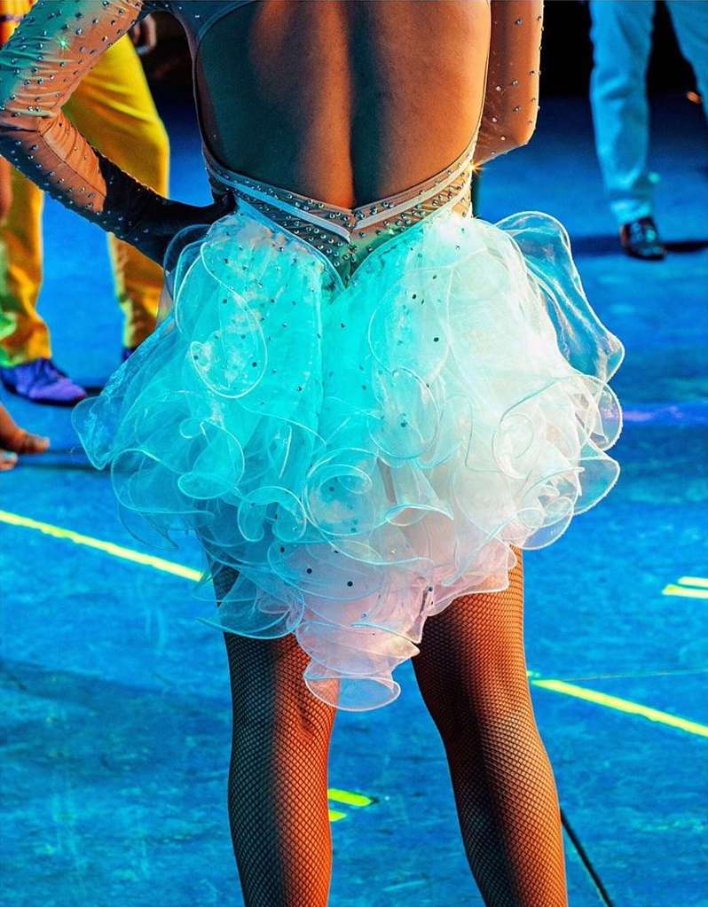 Close-up back view of a female dancer in a white ruffled dress with sparkles, and fishnet stockings on a brightly lit stage.