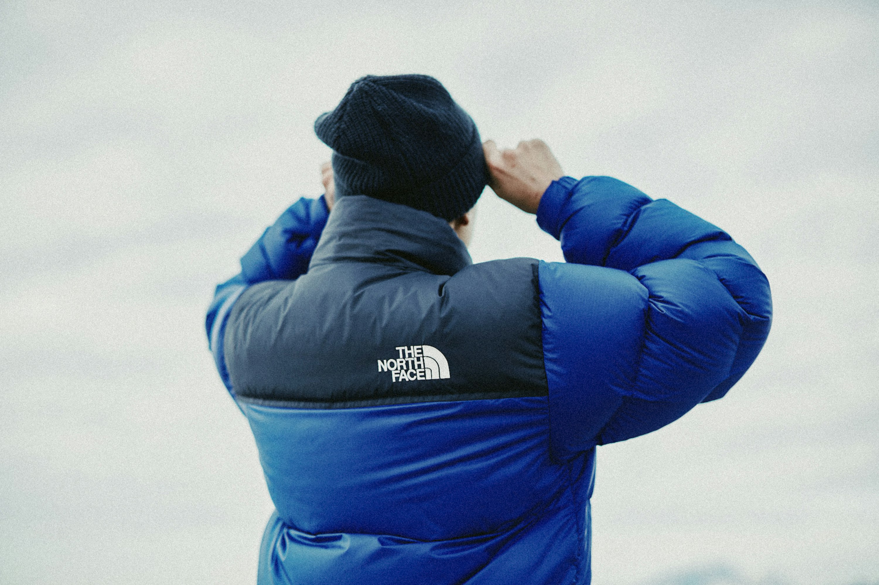 Adult wearing a blue and black The North Face puffer jacket and dark beanie outdoors, adjusting the beanie; overcast sky background.