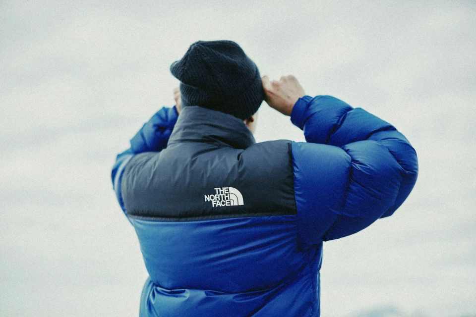 Adult wearing a blue and black The North Face puffer jacket and dark beanie outdoors, adjusting the beanie; overcast sky background.