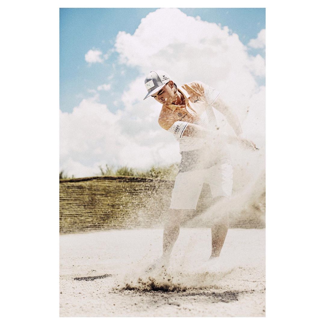 Action shot of a male golfer in mid-swing on a sand bunker, showcasing sportswear, cap, and sandy environment against a bright blue sky.