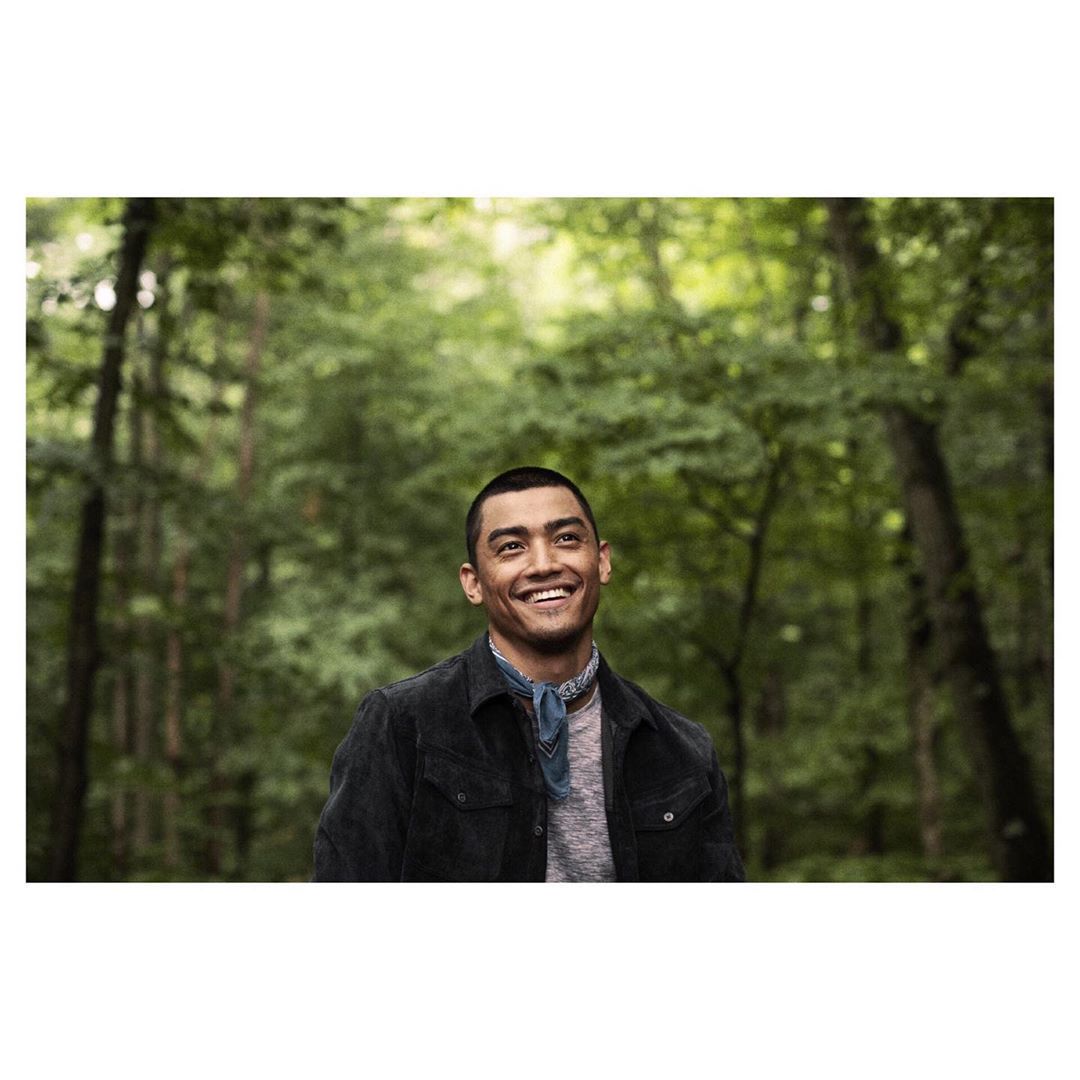 Smiling man in a dark jacket, wearing a patterned bandana, stands outdoors surrounded by tall green trees in a forest setting.