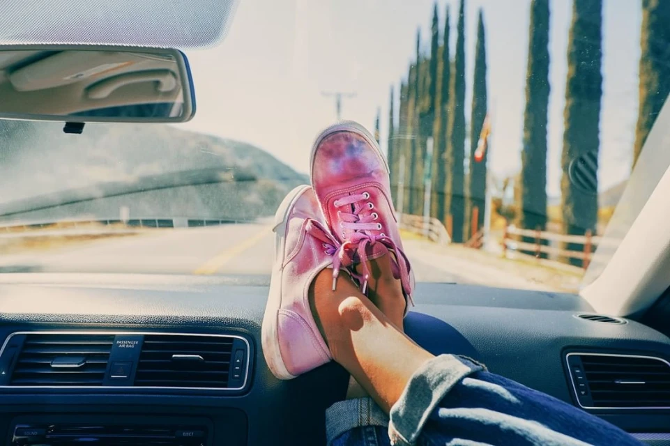 Pink sneakers on car dashboard, legs visible, driving on open road lined with tall trees. Sunny, clear day for a road trip.