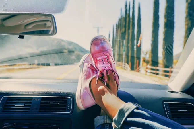 Pink sneakers on car dashboard, legs visible, driving on open road lined with tall trees. Sunny, clear day for a road trip.