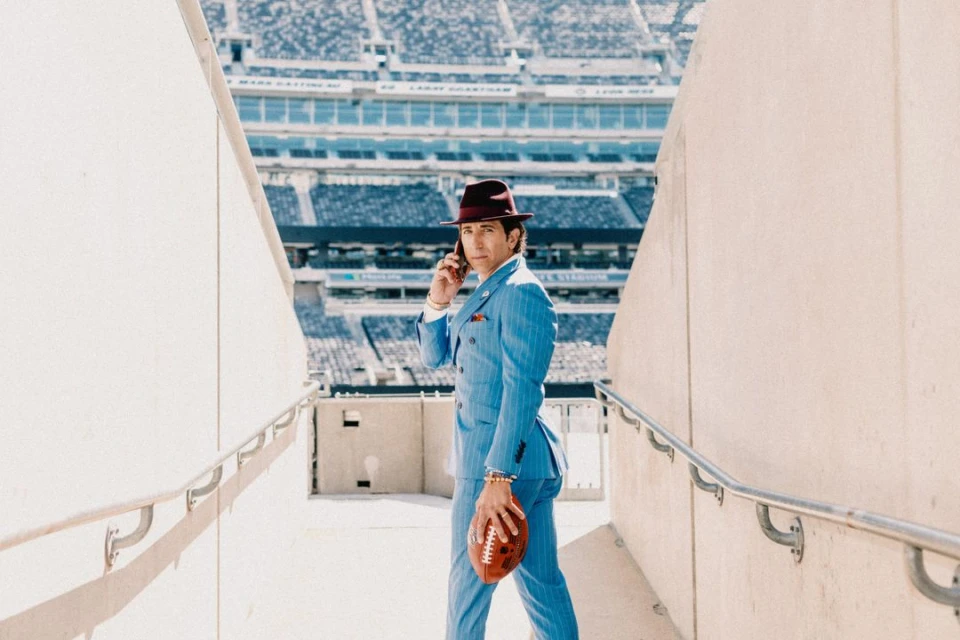 Man in blue suit and hat stands on stadium steps, holding football, talking on phone, with bright sunlight and empty stadium seats.