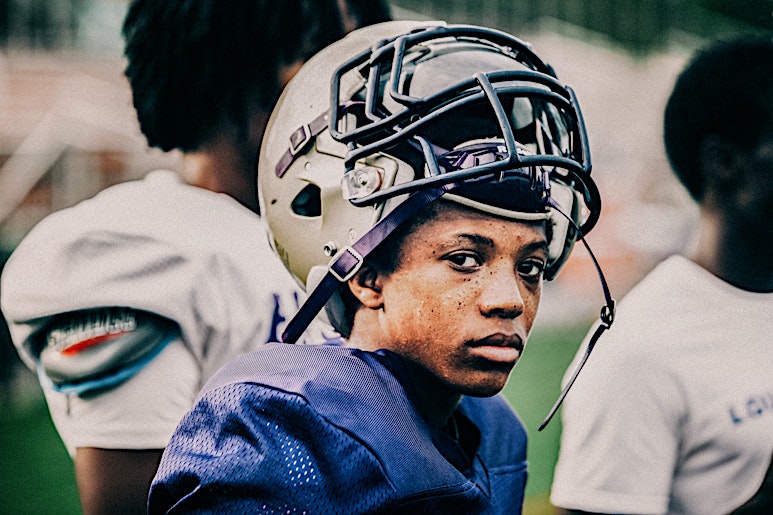 Portrait of a young African American male football player in uniform and helmet, posing outdoors on a grassy sports field with teammates.
