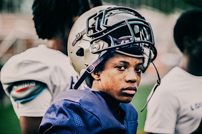 Portrait of a young African American male football player in uniform and helmet, posing outdoors on a grassy sports field with teammates.