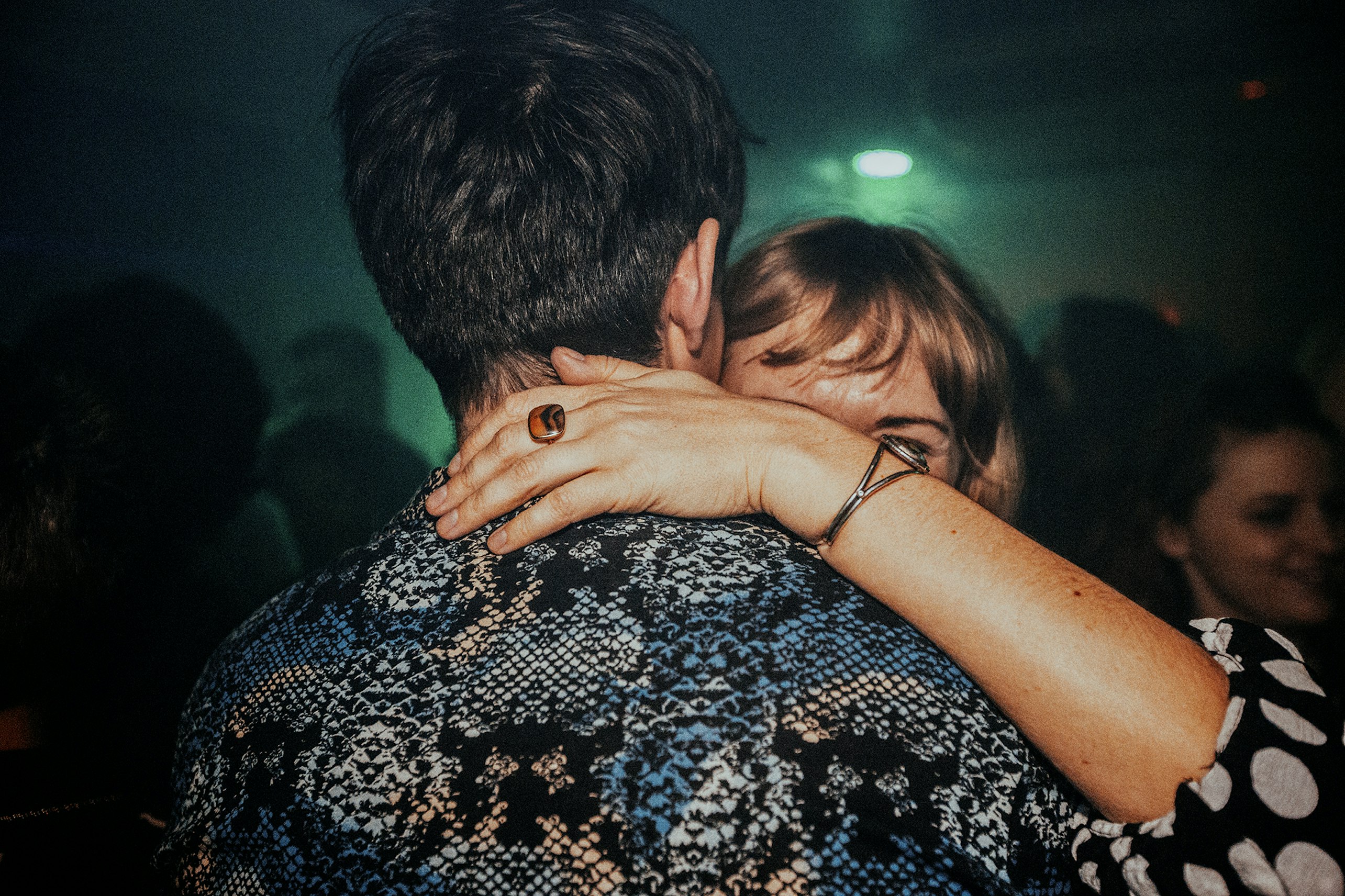 Close-up of a couple embracing in a dimly lit indoor setting, the woman's arm around the man's neck, with visible bracelet and ring.