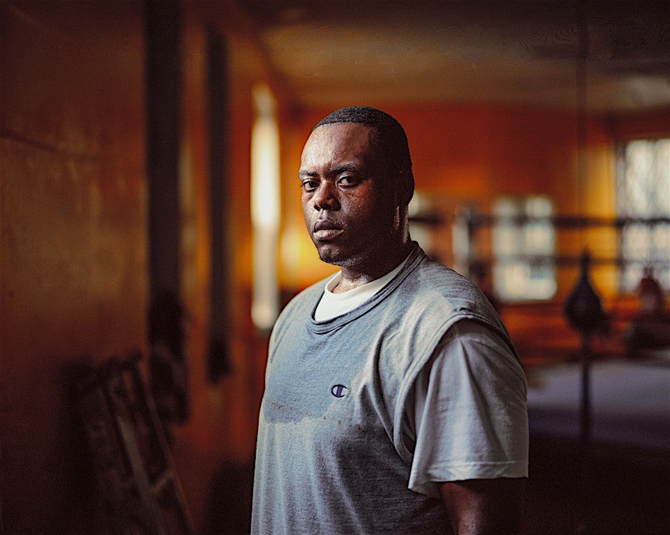 Portrait of an African-American man in a blue Champion shirt, looking at the camera in an orange-toned indoor gym setting.